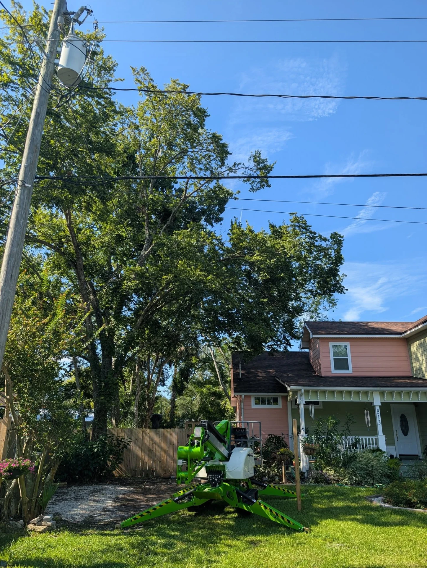 Compact spider lift set up in a front yard to work hardwoods growing into the power lines beside a pink two-story home